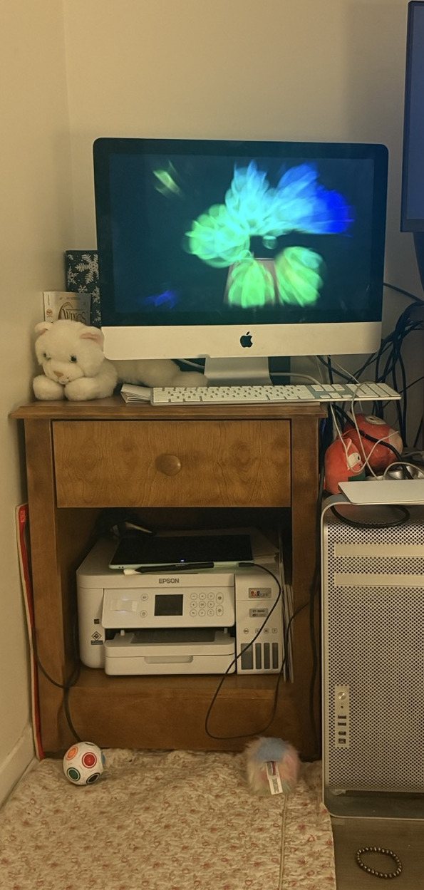 The iMac on smaller dressed in full view. It's packed here, but not stuffy. The keyboard is with number pad; that last third's past the edge. The trackpad sits ontop of the Mac Pro and is attached by wire. In the space beneath the drawer there's an Epson printer and on top of that a Wacom Intuos drawing tablet. A little kitty plushie sprawls comfortably; there are two strawberry plushes on the Mac Pro, too. In this thumbnail the display shows the Arabesque screen saver, but if you click the full image, there's iTunes playing back some music by Phyrnna.
