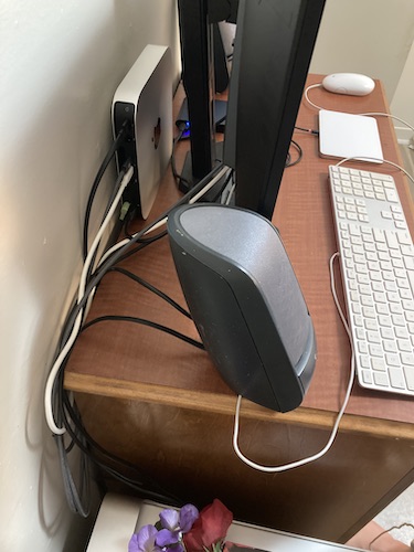 Shot of cable management behind the monitor and speakers, with the Mac Mini standing on the desk, almost flush against the wall; there are a number of wires threading from each device, but almost all find their way behind the desk, leaving very little visible tangle.