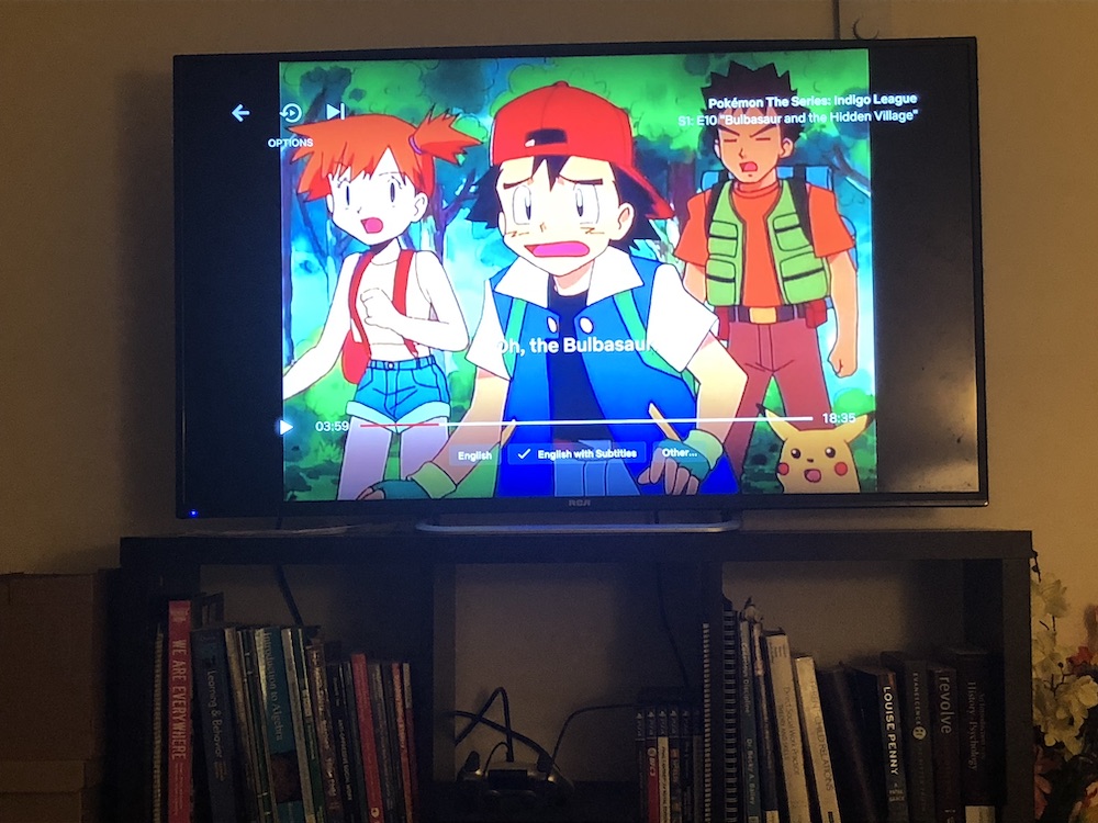 photo of a TV on an ikea quality book-holding stand; Pokémon is playing on Netflix, showing Ash, Pikachu, and their friends making shocked expressions
